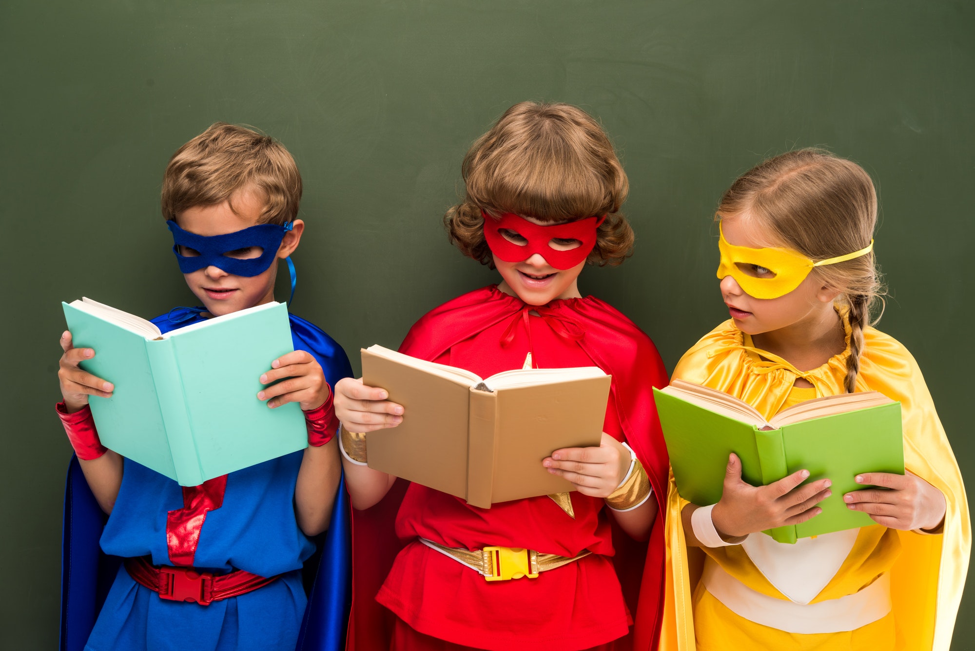 little superheroes in costumes reading books, chalkboard behind